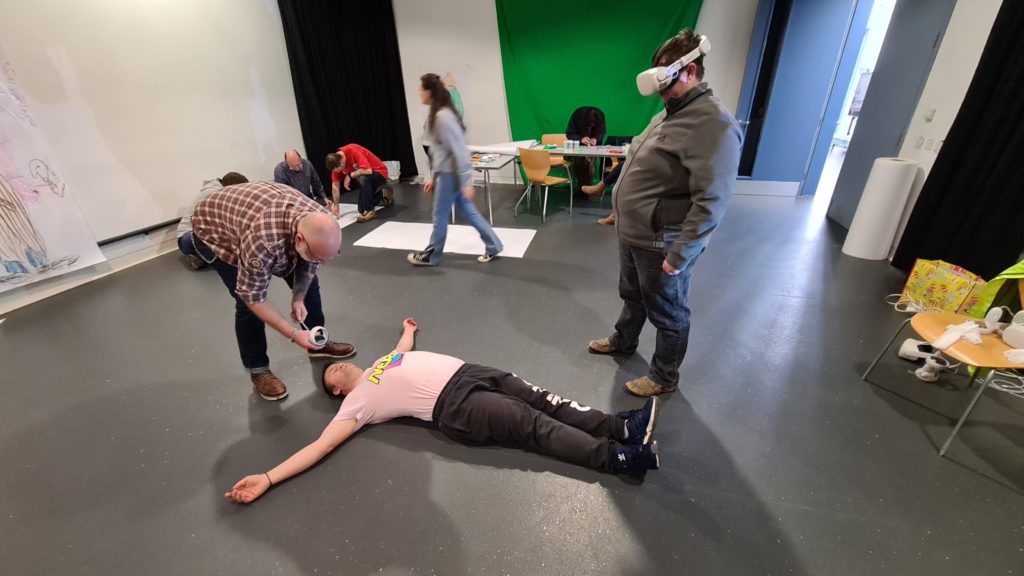 Three male participants viewing and drawing using VR equipment. One participant is laying on the floor being drawn around, while another views in VR.